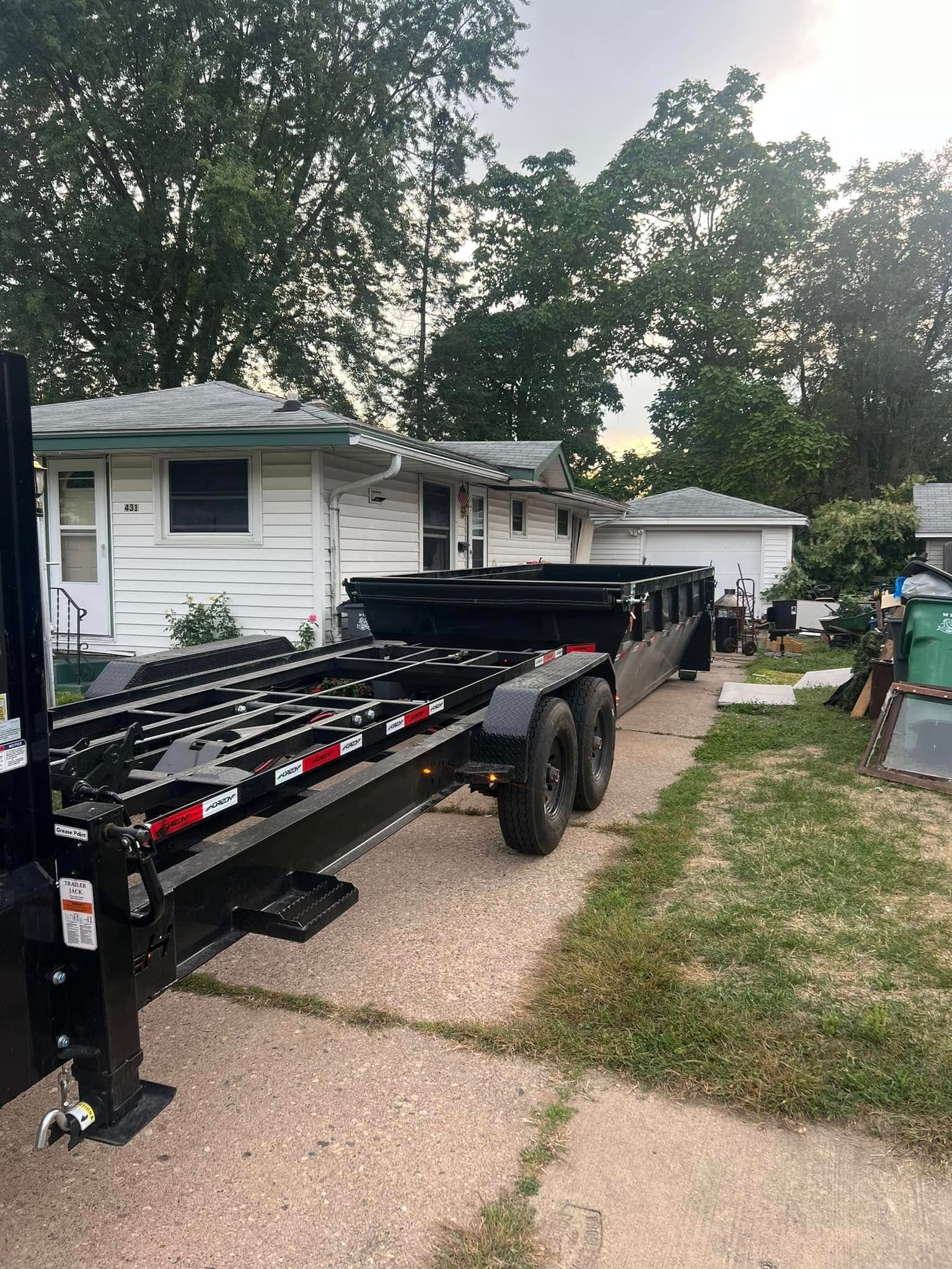 A black flatbed equipment trailer parked on a gravel driveway in front of a white house.