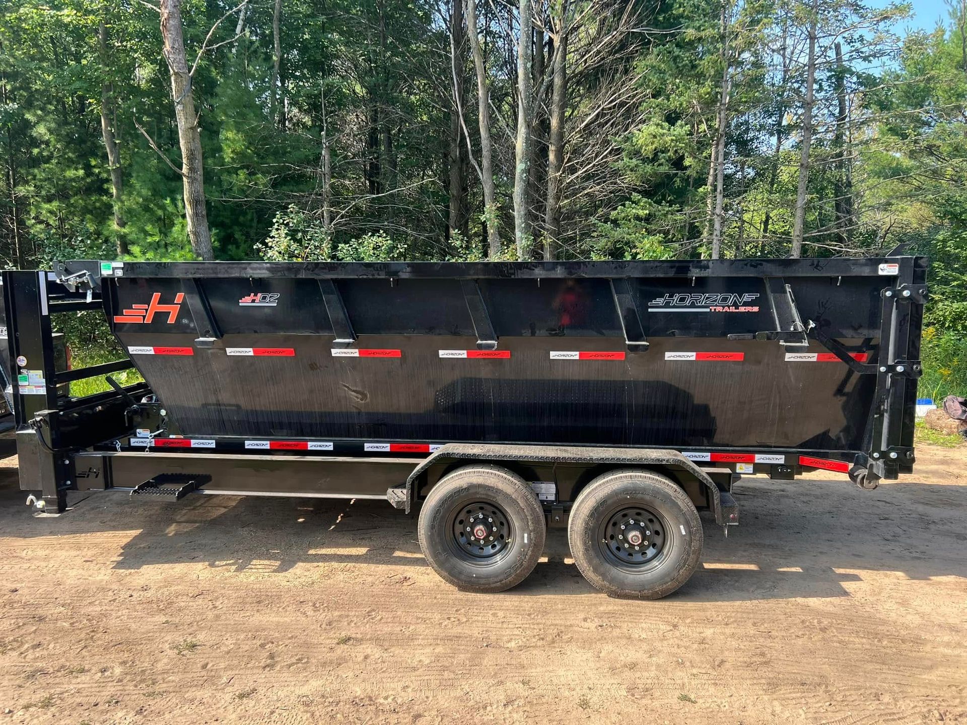 A black dump trailer parked on a dirt lot, featuring orange branding and reflective safety tape along the sides.