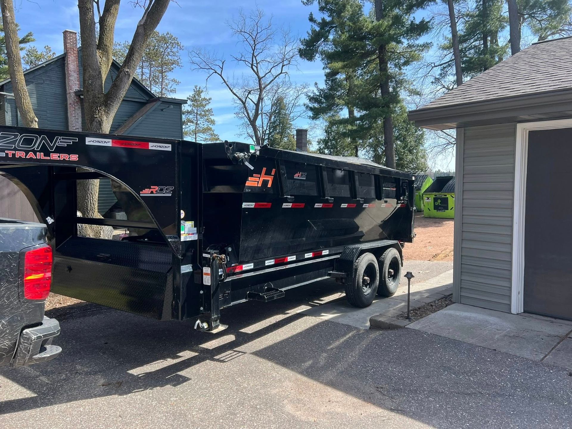 A black dump trailer attached to a pickup truck parked in a driveway next to a building and trees.
