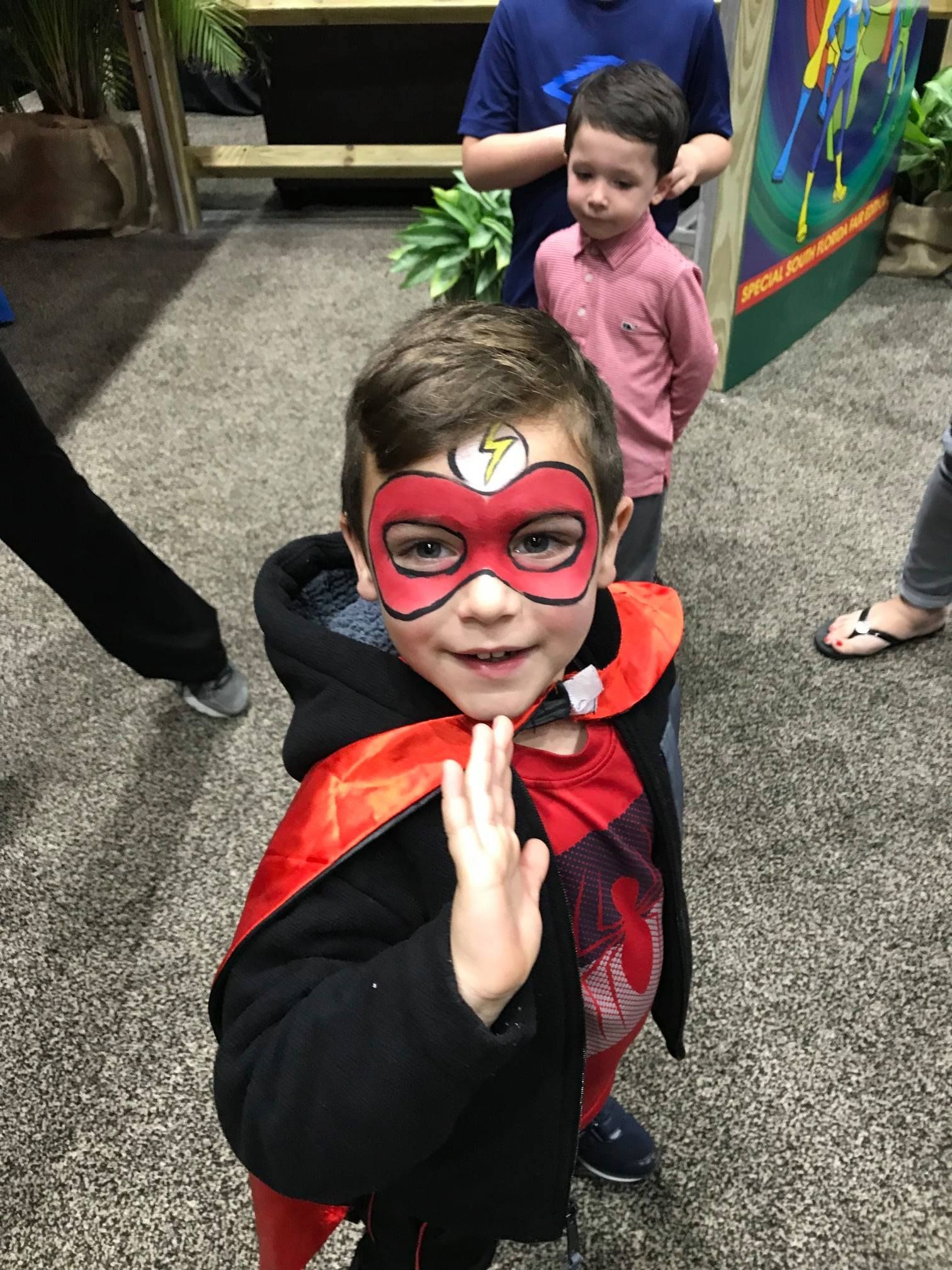 A child wearing a red cape and face paint featuring a superhero mask and lightning bolt emblem, waving at the camera.