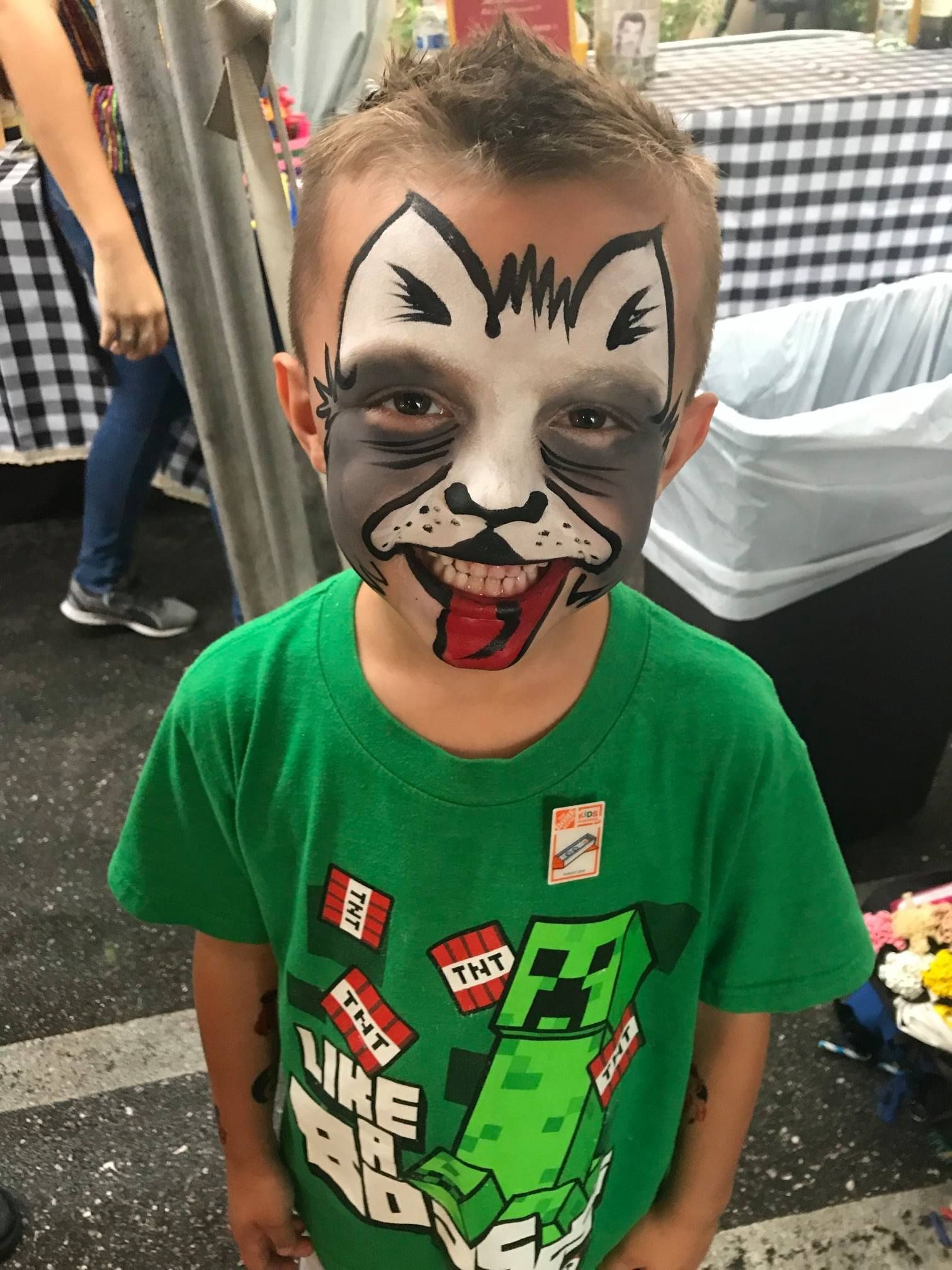 A child with cat face paint, including a black-and-white muzzle and a red tongue, wears a green Minecraft-themed T-shirt.