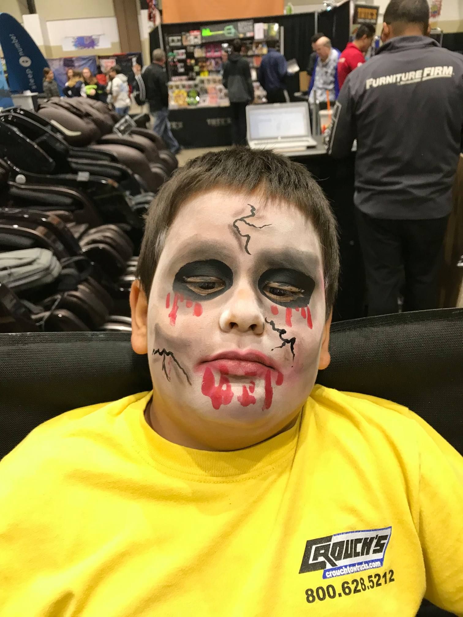 A person wearing yellow, with zombie-themed face paint, seated in an indoor event space with booths and people in the back.