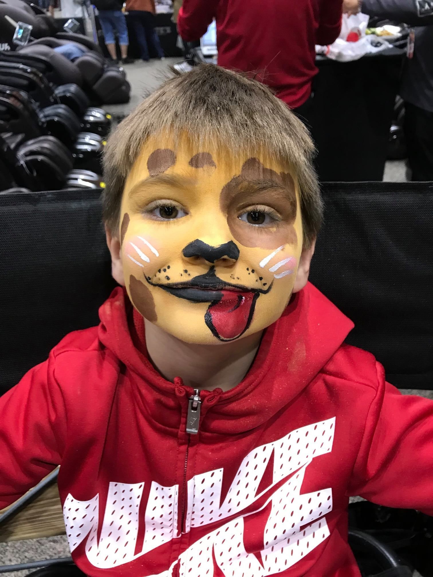 A child wears yellow, brown, and black face paint to look like a dog, wearing a red hooded sweatshirt in a store.