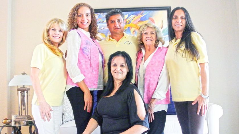 A group of six people posing together indoors, wearing yellow shirts and pink vests, smiling at the camera.