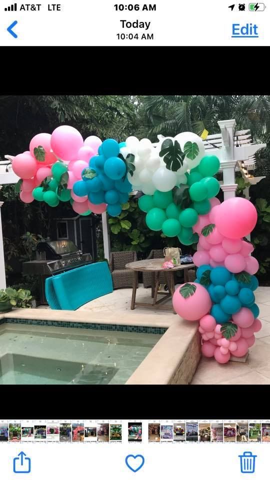 A vibrant balloon arch featuring pink, teal, and white balloons accented with tropical leaves by a backyard pool.