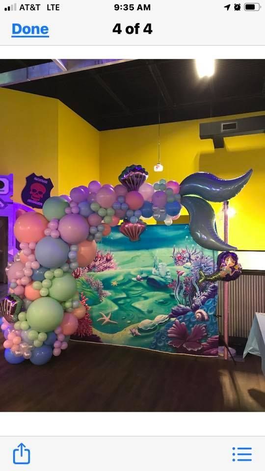 A colorful balloon arch with a mermaid tail foil balloon decorating a mermaid-themed backdrop in an indoor room.