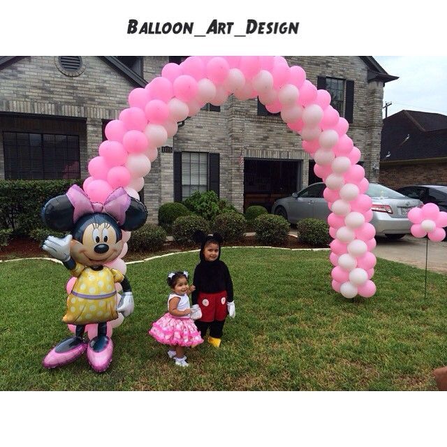 A pink and white balloon arch stands in a front yard, next to a Minnie Mouse balloon and two children in costumes.