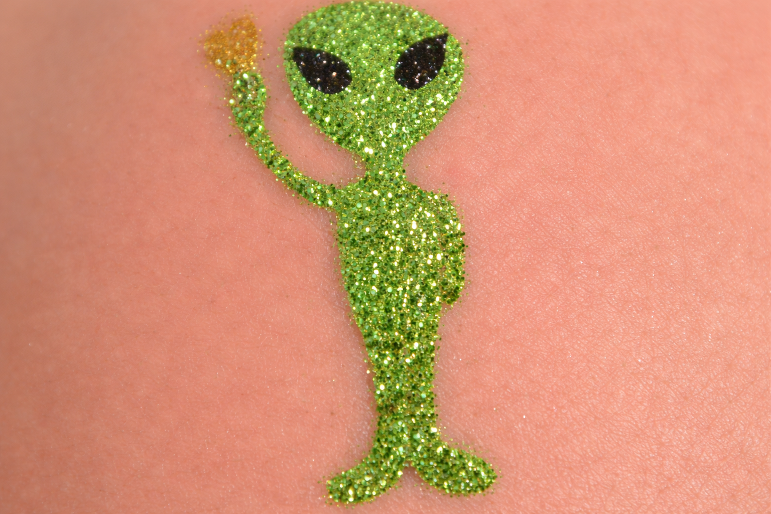 A glittery, green alien sticker against a peach-colored background, waving one hand while holding a small yellow heart.