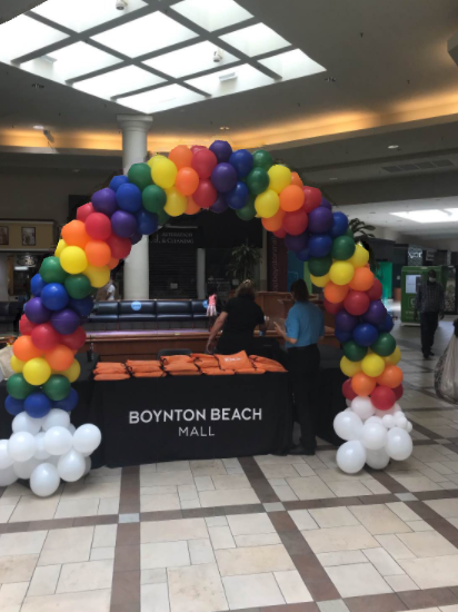 A rainbow balloon arch over a Boynton Beach Mall information table with white balloon clouds at the base.
