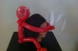 A red balloon figure sits on a person's head, with its arms wrapped around the sides and holding white balloon shapes.