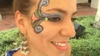 Close-up of a person with decorative face paint and a rhinestone near the eye, smiling outdoors.