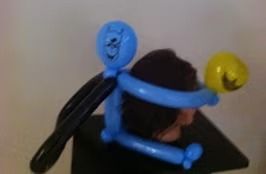 A human head wearing a balloon structure shaped like a blue figure with black wings and a yellow ball held in its arms.