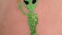 A green glitter alien tattoo on skin, shown with an arm raised in a wave.