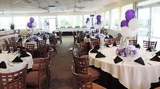 Event venue with round tables set for dining, featuring white tablecloths, black napkins, and purple and white balloons.