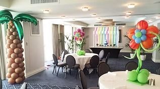 Event space decorated with balloon sculptures of a palm tree and a large flower, featuring round tables and chairs.