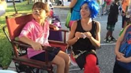 A child in a pink shirt sits in a red folding chair beside a person with blue hair wearing a black top and red leggings.