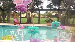 Balloon decorations shaped like flowers in purple, pink, and teal, set up on a table beside a backyard swimming pool.