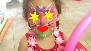 A child with cat face paint, a pink flower lei, and a red top, holding a pink balloon at a party.