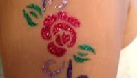 A sparkly glitter tattoo on skin featuring a bright red rose with green leaves and purple accents.