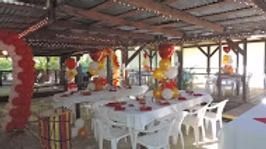 An outdoor pavilion decorated with red, yellow, and white balloons, featuring banquet tables set for a celebration.