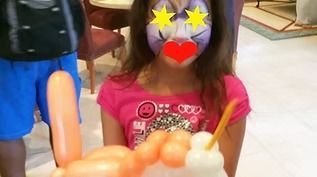 A child with face paint and a pink shirt holds an orange and white balloon sculpture in an indoor setting.