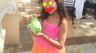 A person in a bright pink and orange dress holds a green, decorated toy outdoors on a paved patio.