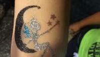 A glittery temporary tattoo of a fairy sitting on a crescent moon with small stars nearby on skin.