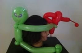 A green balloon alien figure sits on a person's head, aiming a red balloon ray gun.