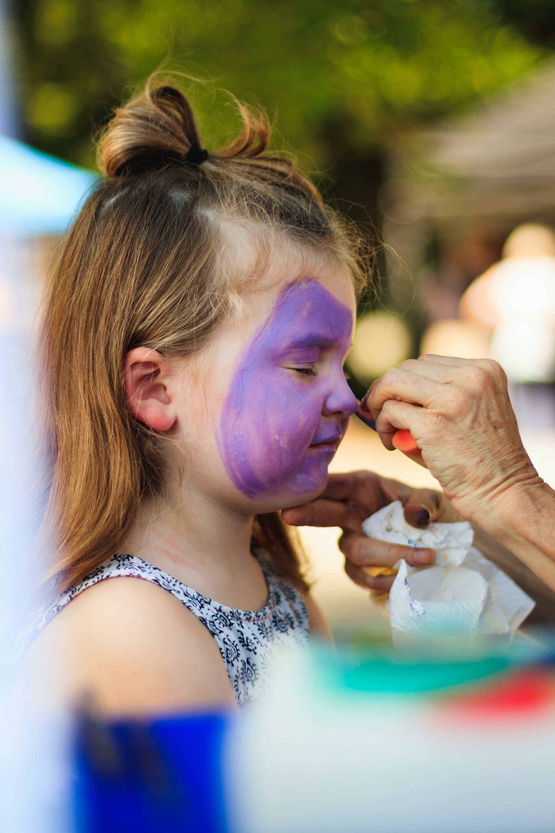 A person applying purple face paint to a young person’s nose in an outdoor setting.