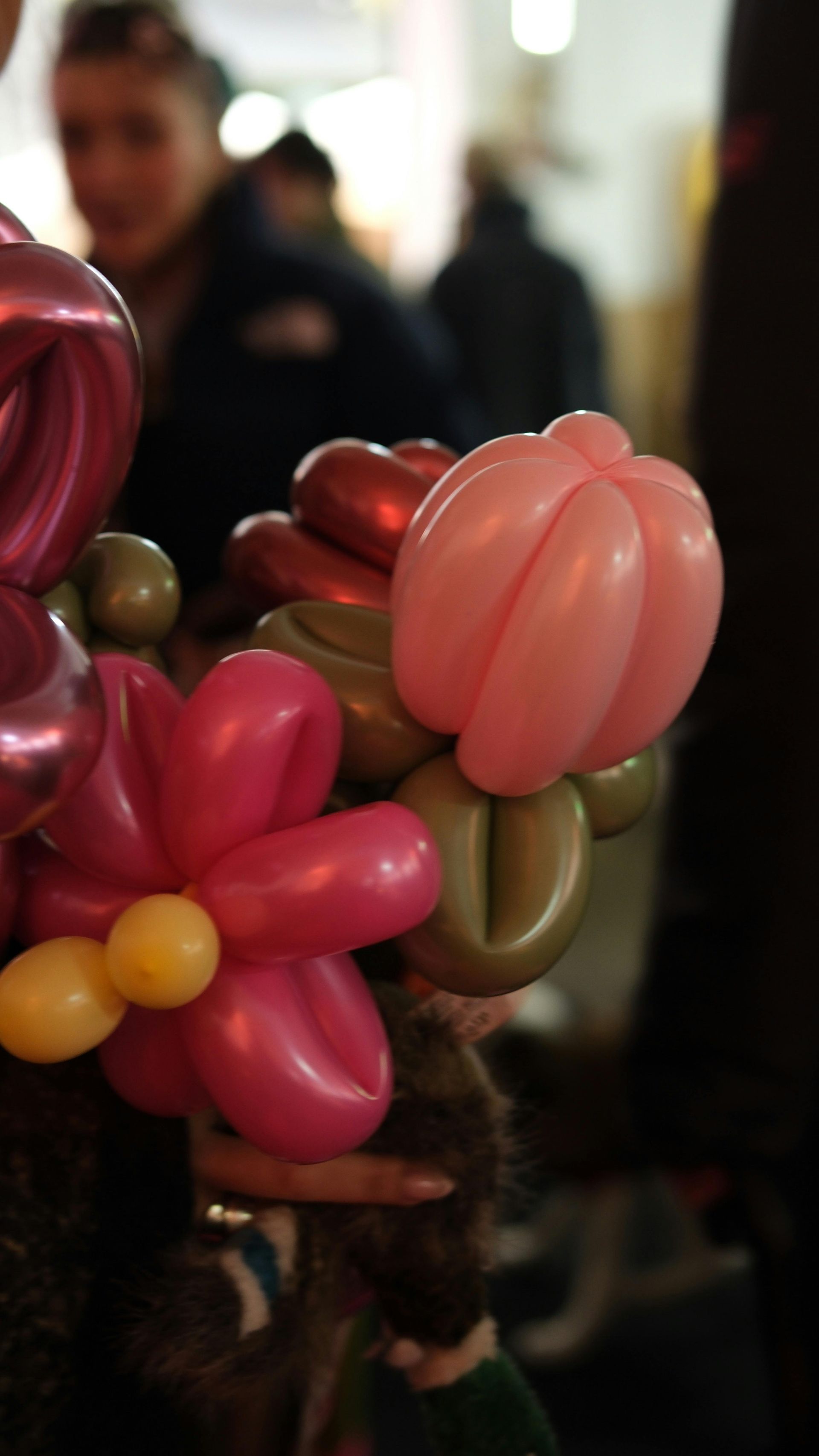 A hand holds a colorful balloon sculpture featuring a pink flower with yellow centers and a light pink spherical balloon.