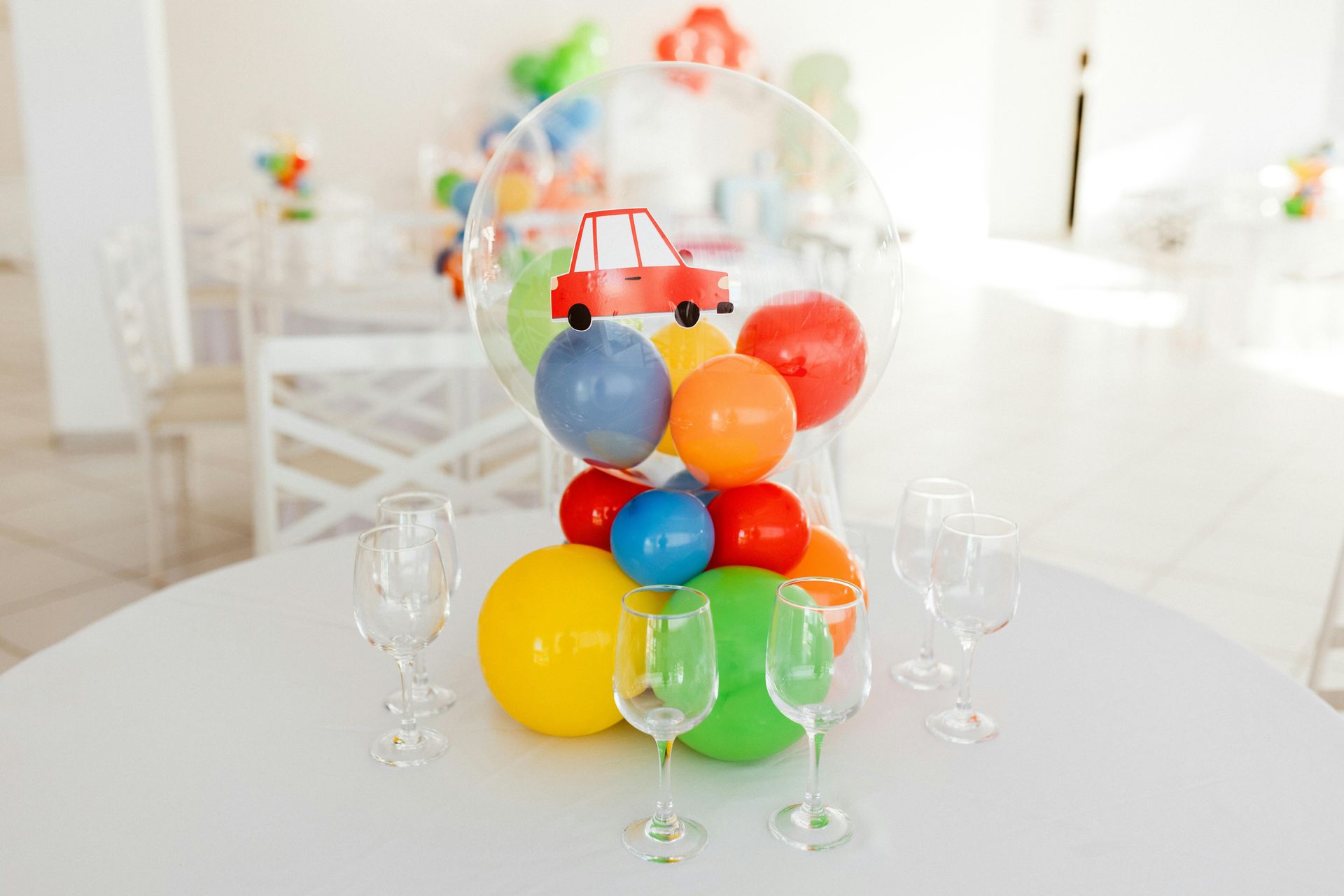 A balloon centerpiece featuring a small red car inside a clear bubble balloon, surrounded by colorful balloons and glasses.