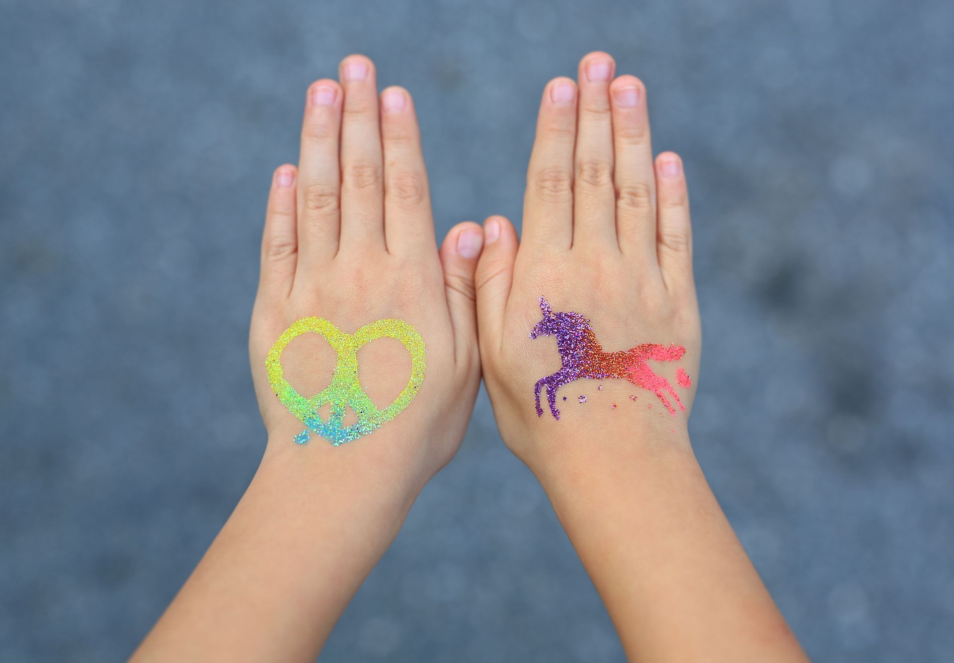Two hands side-by-side with a glittery green peace sign on the left and a multicolored glitter unicorn on the right.