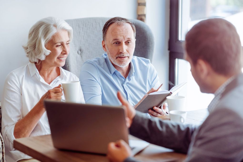Three people in a meeting, one showing a tablet while others listen with a laptop open.