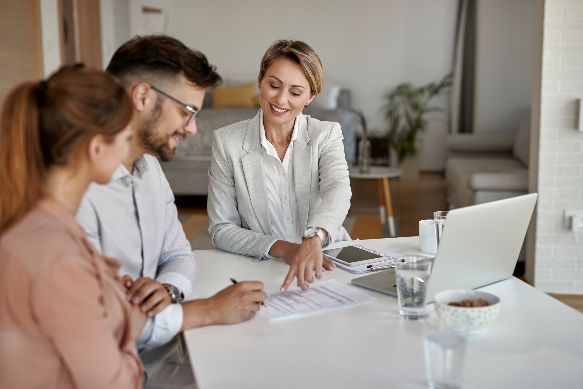 Three coworkers review documents at a white table in a bright office, with a laptop and coffee nearby.