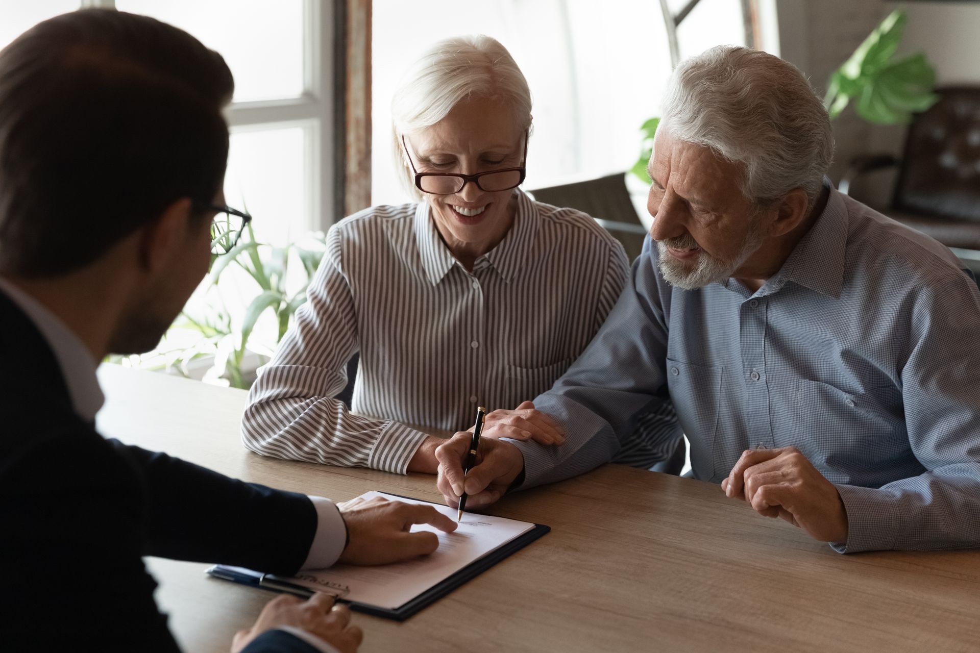 Three people discuss paperwork at a table in a bright office, with one person taking notes. Three people discuss paperwork at a table in a bright office, with one person taking notes.