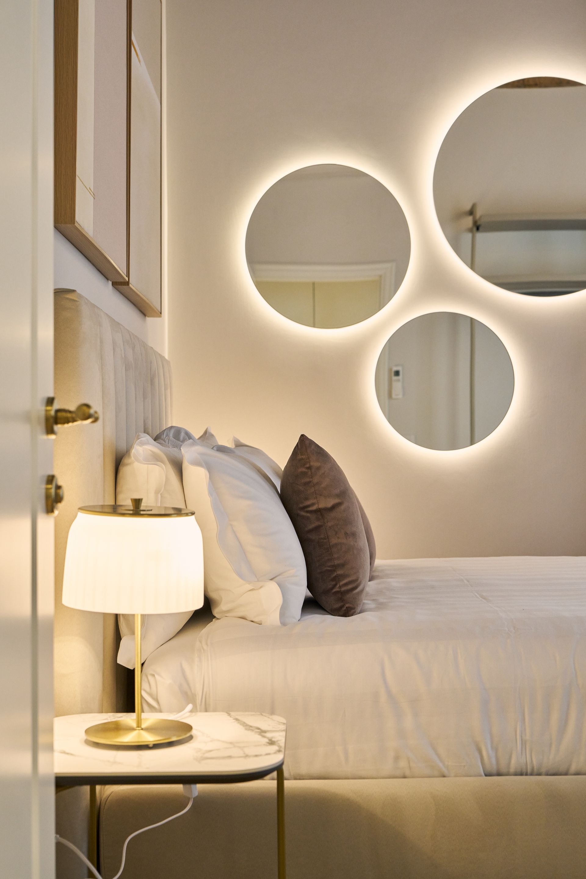 Cozy bedroom with a neatly made bed, gold lamp, and round backlit mirrors on a beige wall