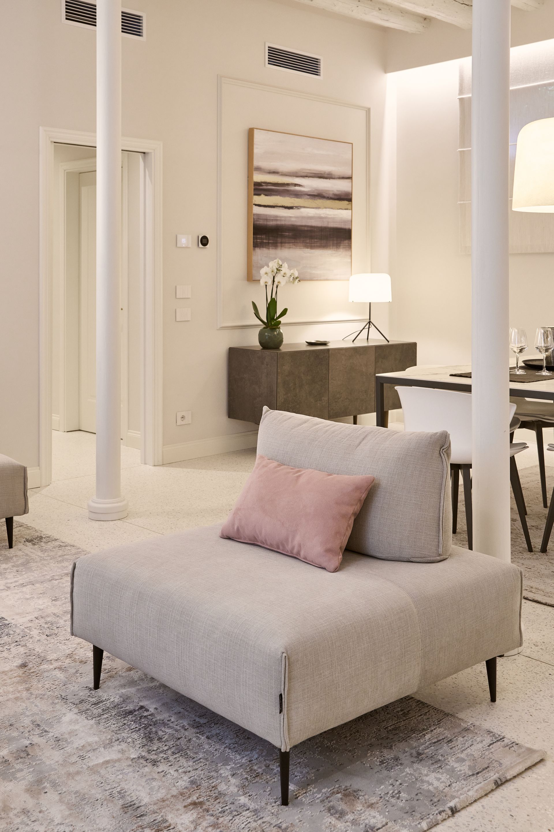 Modern living room with a gray chaise, pink pillow, rug, and a lamp beside a dining area.