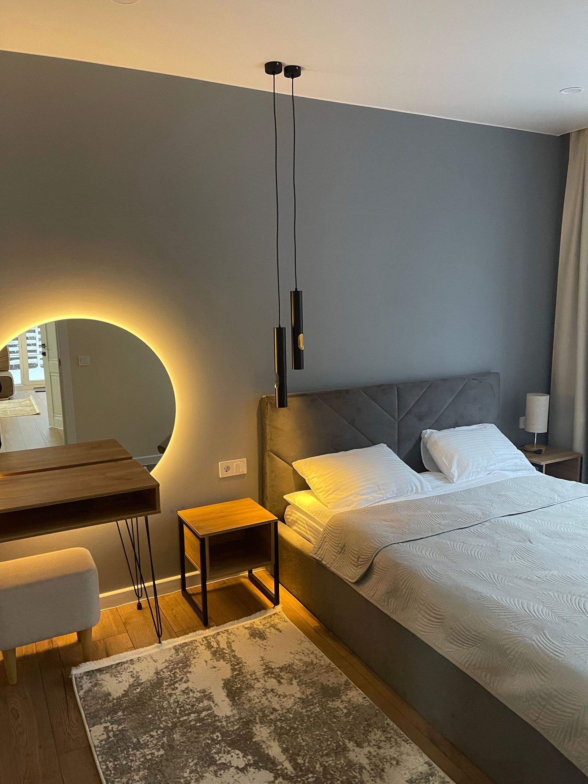 Modern bedroom with gray bed, glowing round mirror, wooden nightstand, and warm lighting.