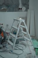 Paint-covered step ladder in a dusty room with tools, wires, and boards during renovation