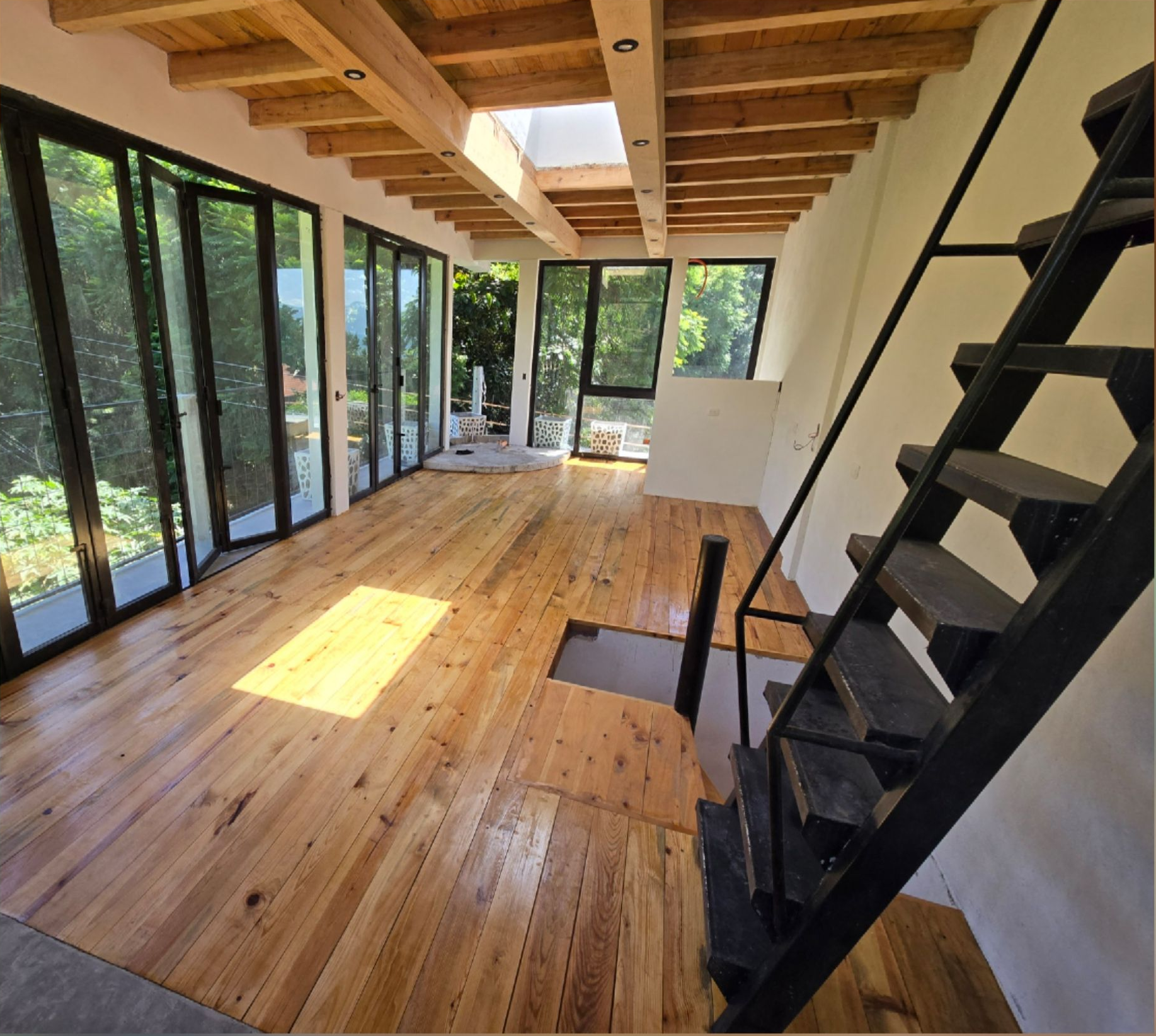 Bright sunroom with wood floors, exposed beams, floor-to-ceiling windows, and black staircase.