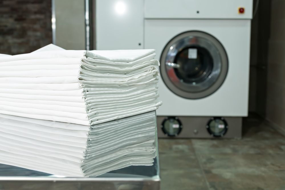 Pile of Folded Linens In Front Of Washing Machines In A Laundromat — Dapto Swish & Swash Laundrette In Dapto, NSW