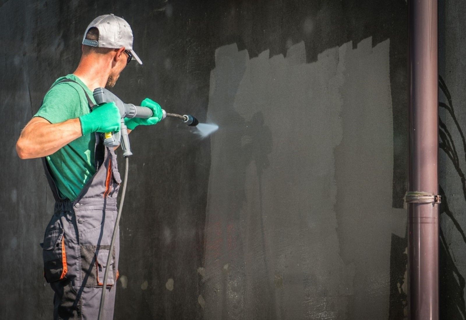 Worker spray-painting a dark wall with a green paint sprayer, wearing gloves and overalls.