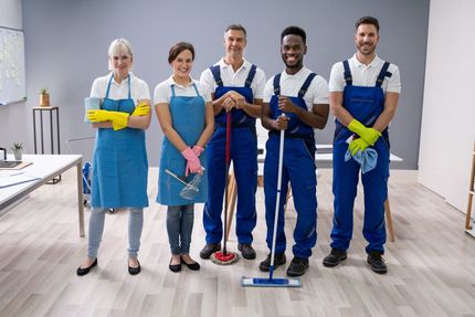 Five cleaners in blue uniforms stand with mops and cleaning supplies in a bright room.