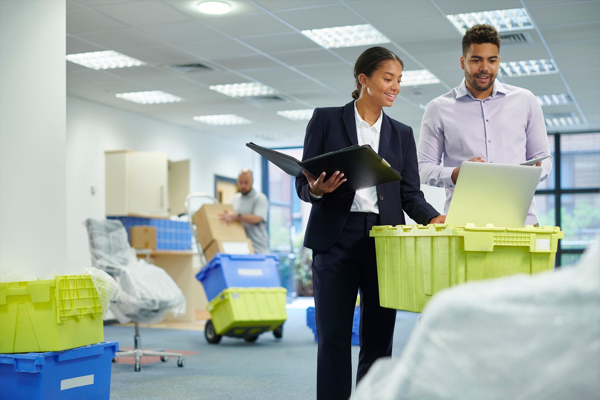 Two coworkers with a laptop in a bright warehouse, surrounded by stacked boxes and carts.