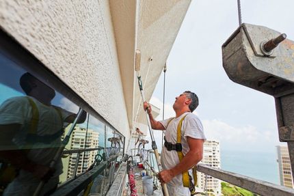 Worker on a high-rise scaffold cleaning a building facade with a squeegee, ocean in the background