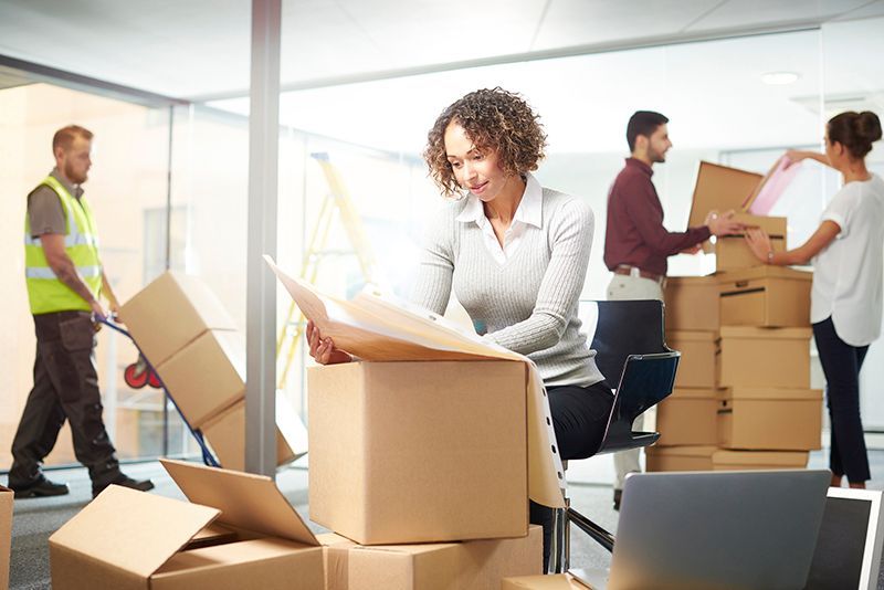 People moving boxes in a bright office during a move, with one woman unpacking at a desk.