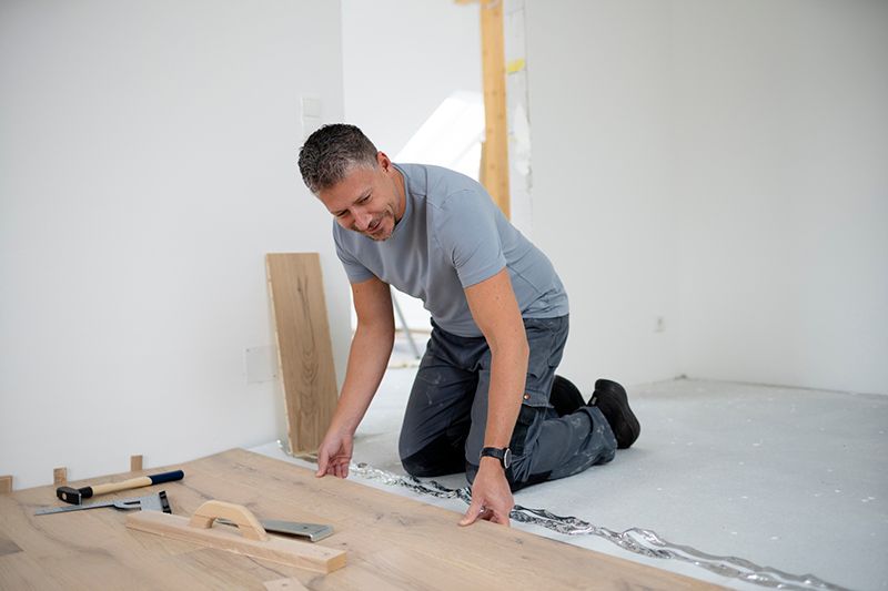 Man kneeling on a floor, measuring and installing wooden flooring in a bright room.