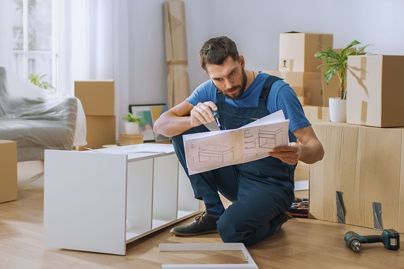 Man assembling a white cabinet in a bright room, reading instructions beside moving boxes and tools.