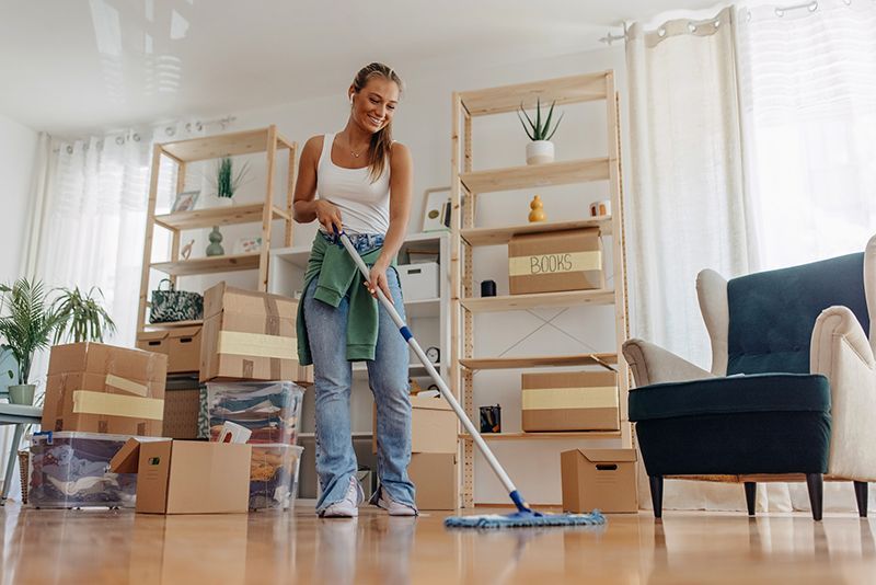 Woman mopping a wooden floor in a bright living room with moving boxes and furniture.