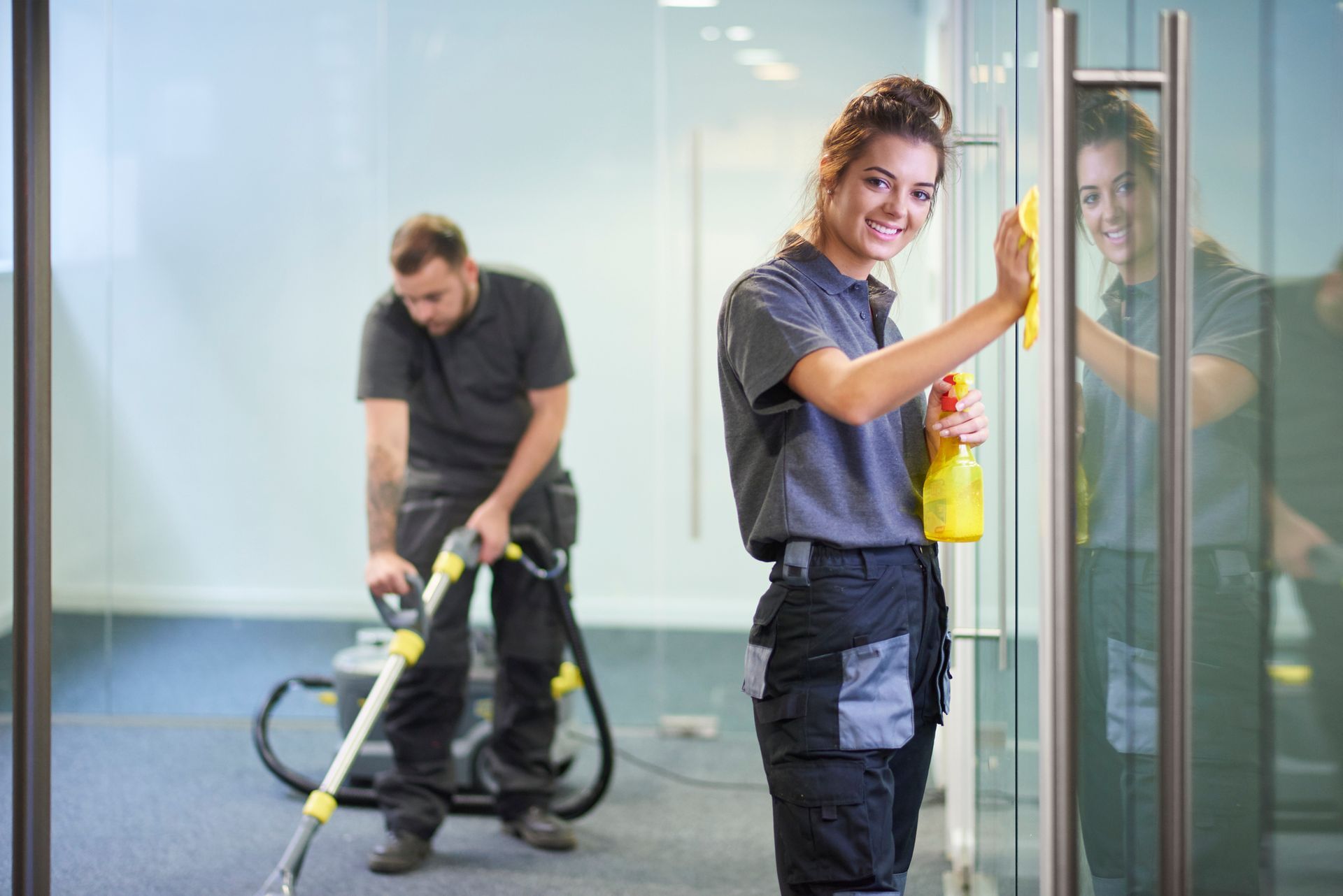 Two cleaners working in a glass office, one wiping a glass door and one vacuuming the floor.